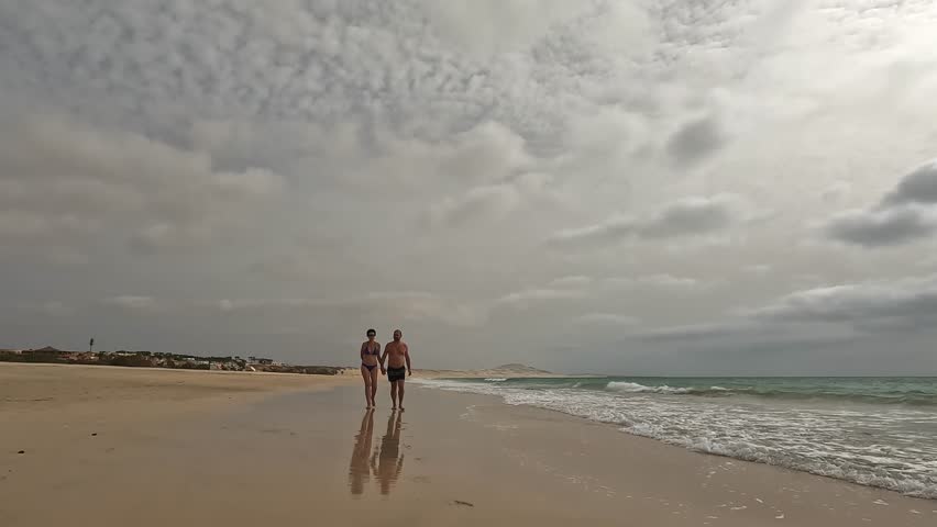 Romantic couple walks hand in hand along peaceful, sandy beach of Boa Vista island with cloudy sky overhead, creating serene and intimate moment by the ocean, Cape Verde. Slow motion