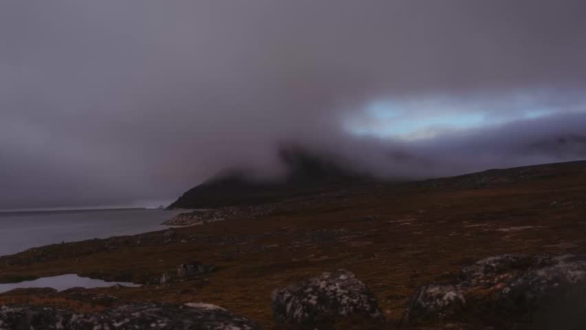 Timelapse of a cloudy day in Greenland, showing fog rolling over a beach toward the water, with occasional sunlight breaking through and icebergs floating in the sea.