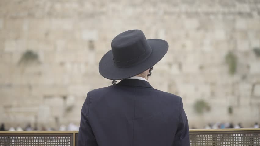 Jewish man praying at the Western Wall in Jerusalem, Israel. He has long side curls and wears a black hat and coat. Camera moves in to subject in slow-motion with selective focus. From back view.