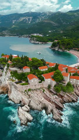 Island of Sveti Stefan - Montenegro. Aerial view. Drone going backwards. Waves breaking on the rock. Famous and luxury travel destination. Boats in the bay. Turquoise water.