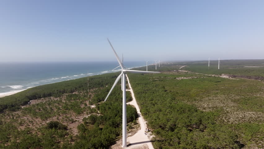 Rotating wind turbine stands near a sandy beach, showcasing the ocean coastline and green trees under a blue sky