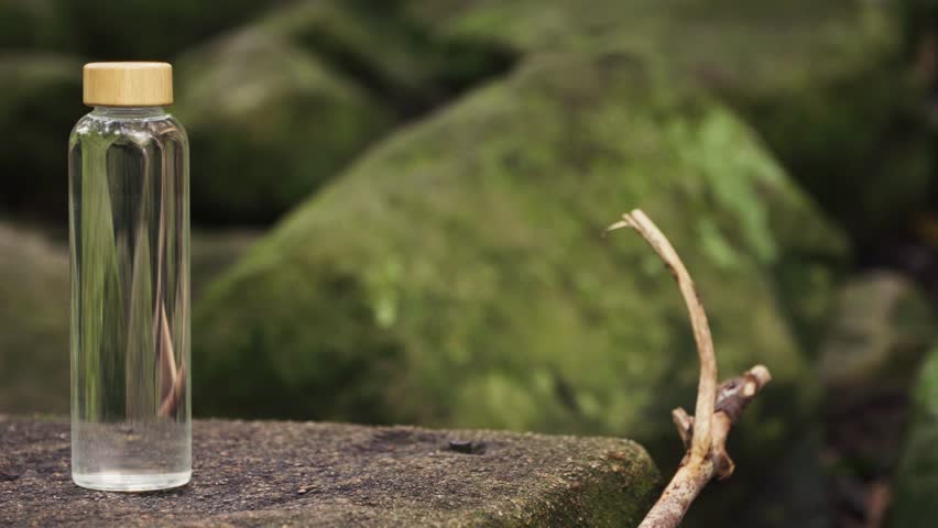 Close up, Pan left, A glass water bottle sitting on a rock with green moss covered rocks and wet logs and branches in the background.