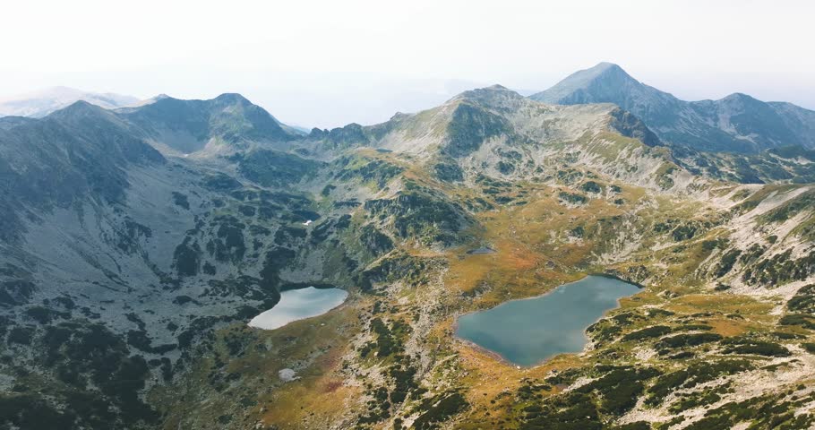 Multiple glacial lakes spread across vibrant green pastures surrounded by tall mountain ranges, in the sun light, tracking drone shot