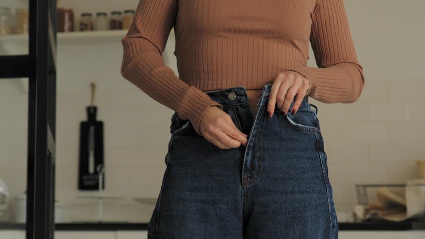 Close-up of overweight attractive mature woman trying to zip small pants after gaining weight while standing in domestic room. Midsection.