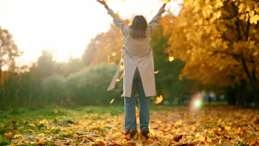 Blonde woman in trench coat enjoying fall season in forest, throwing foliage up . Happiness and joy, carefree female lady admiring beautiful golden trees and breathing cold fresh air, rear view