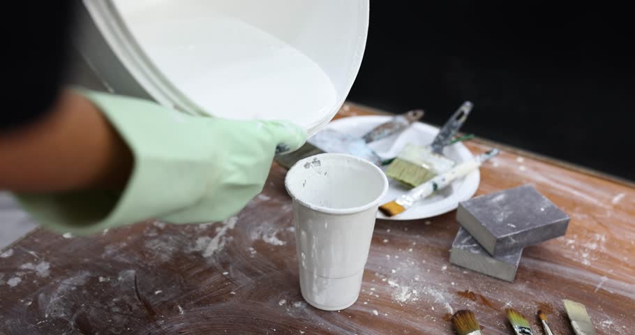 Repairman pours white paint from bucket into plastic cup on dirty table. Professional handyman prepares brushes and paint for painting process