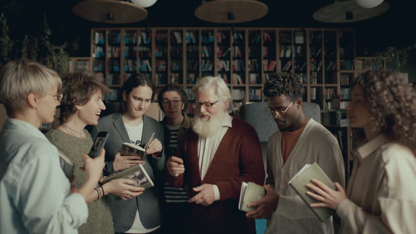 Group of young enthusiastic fans smiling and taking photos with elderly author during book signing event in public library