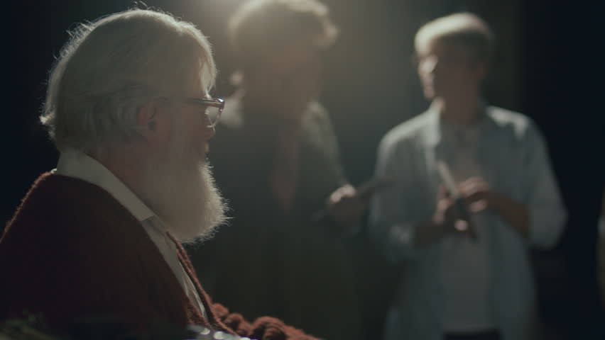Elderly author with white beard and glasses sitting on stage under soft lighting, signing book for reader during launch event