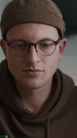 Vertical of Caucasian hipster man dressed in glasses and beanie hat sitting on couch in apartment, looking intently on smartphone screen while watching sports match or checking bet