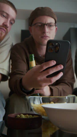 Vertical of sports lovers gambling or watching online match on smartphone while getting together in apartment at weekend, relaxing on sofa, drinking beer and cheering for their favorite team