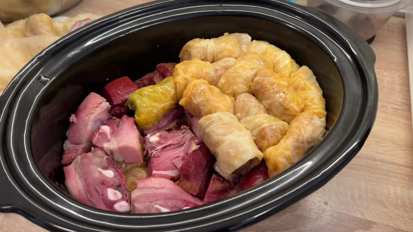 A slow cooker filled with stuffed cabbage rolls and chunks of smoked meat, preparing a Sarma, traditional Serbian food. The cabbage rolls are neatly arranged next to the tender meat pieces.