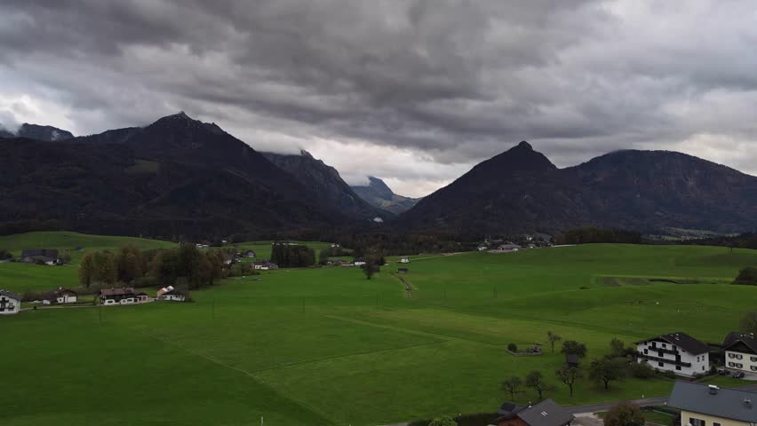 A serene rural landscape featuring a wide expanse of green fields, farmhouses, with dramatic storm clouds gathering over the distant mountain range. Flying over the green field video.