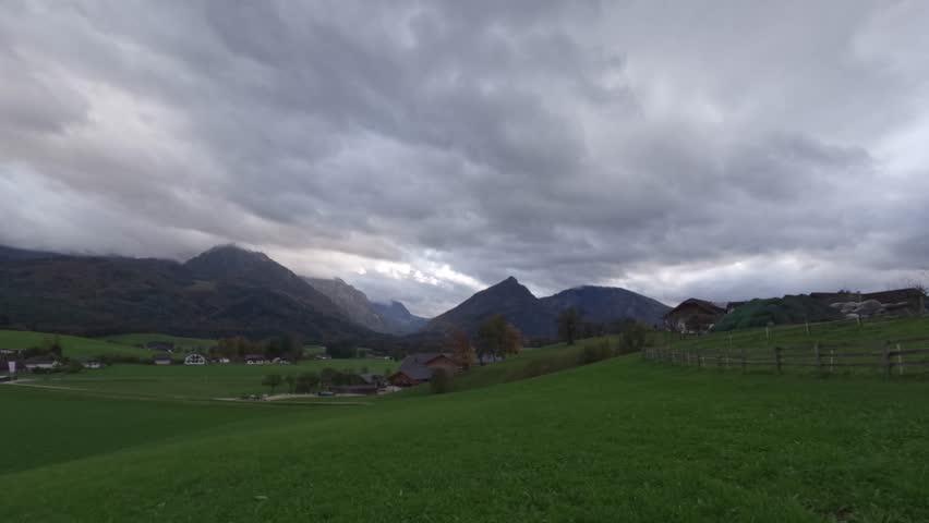 A serene rural landscape featuring a wide expanse of green fields, farmhouses, and wooden fences, with dramatic storm clouds gathering over the distant mountain range. Moving clouds time lapse.