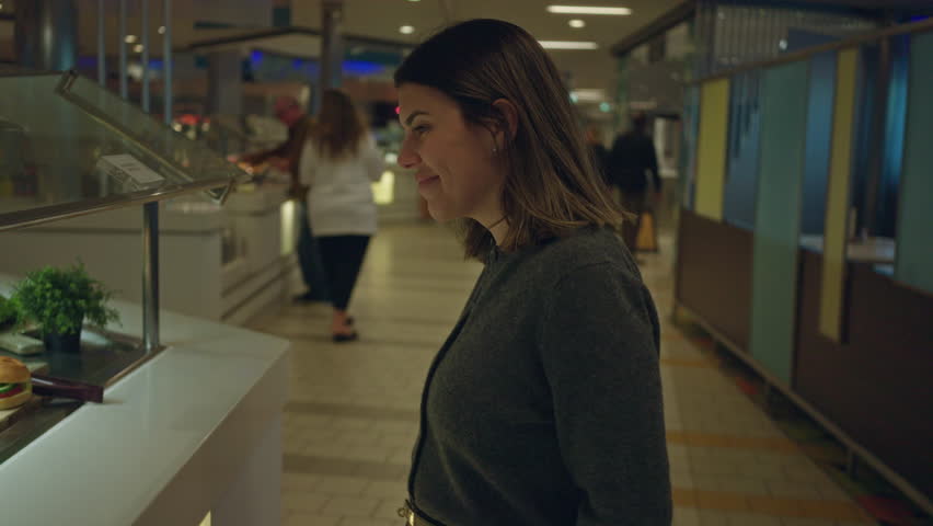 Woman smiling at fresh sandwiches at an indoor buffet restaurant displaying an inviting selection of food while people move in the background.