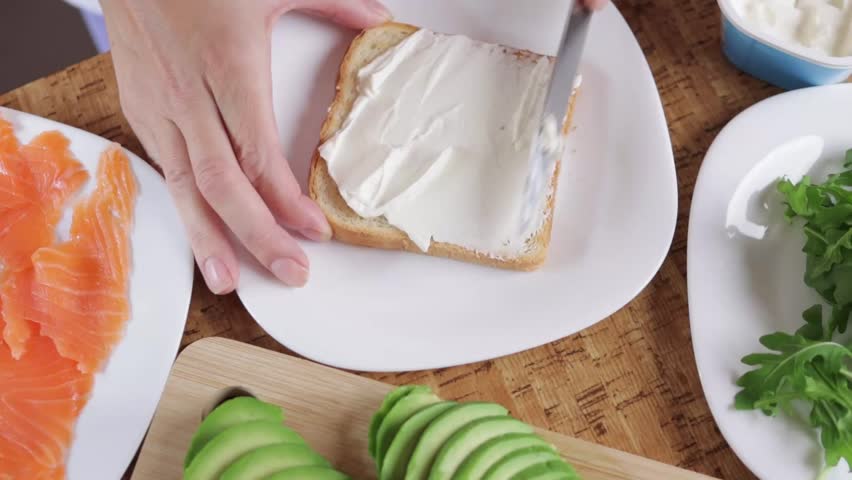 Chef spreads cream cheese on toast, preparing a healthy breakfast or snack. Sliced avocado and smoked salmon await nearby