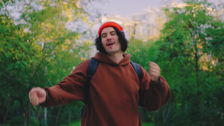 A cheerful man in a red beanie and brown hoodie strolls through a vibrant park. Listening to music, he dances joyfully, blending with the energy of nature
