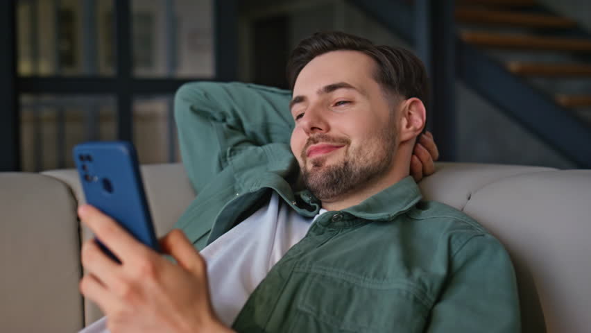 Closeup guy messaging smartphone lying at light couch. Relaxed happy man typing message in social media watching funny content online at weekend. Smiling freelancer using mobile phone at living room