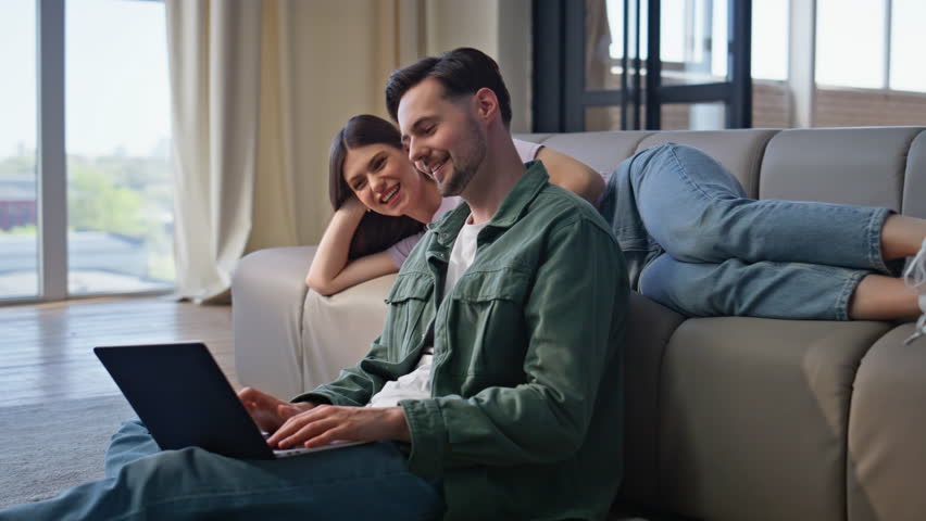 Smiling man working laptop lying girlfriend resting at sofa talking to husband. Couple spending time at home together. Relaxed woman watching computer screen positive boyfriend solving work tasks 