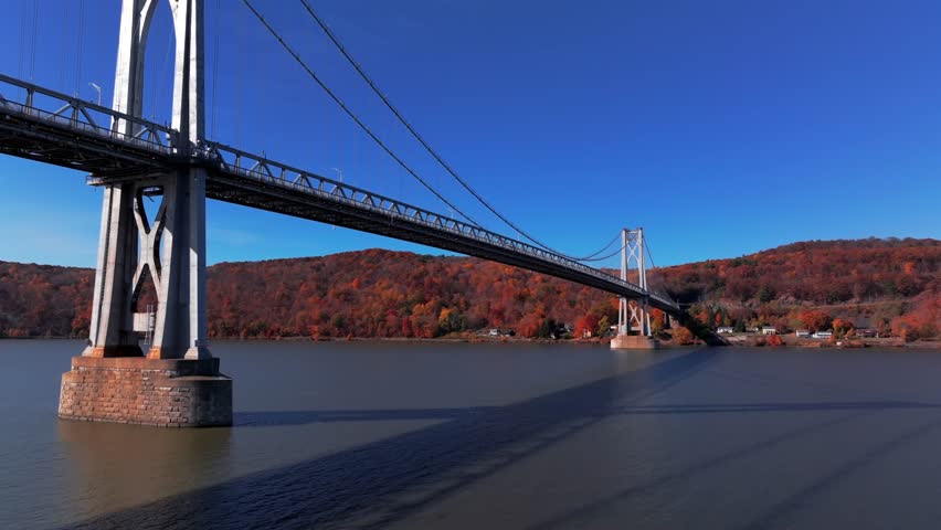 A drone view over the Hudson River by the mid Hudson Bridge on a sunny day during the colorful fall season. The camera dolly in towards and below the bridge span then pan left showing the scenery.