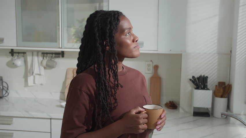 Portrait of positive African American young woman holding cup and smiling for camera in kitchen at weekend