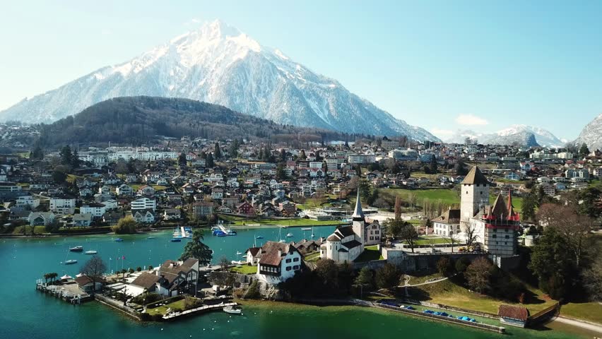 drone pull out shot of Spiez town, castle and church on lake Thun with snow capped niesen mountain in background, showcasing beauty of bernese oberland, switzerland