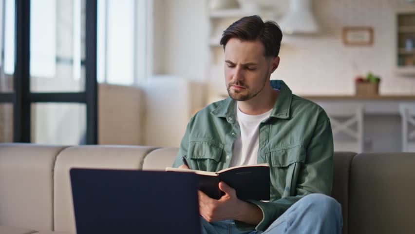 Creative freelancer contemplating solution working remotely at home closeup. Thoughtful man writer looking laptop writing article idea in notebook. Calm pensive businessman thinking project problem 