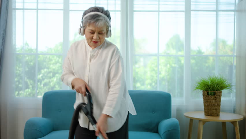 Happy Asian elderly woman using vacuum cleaner to clean living room. Cheerful mature senior housewife doing housework at home. Active retirement grandma vacuuming floor with vacuum cleaner.