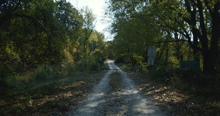 Walking on a path with leaves in a green forest at daytime, a bridge in front, sun is shining, POV, moving forward. Tracking Shot. Kansas, USA.