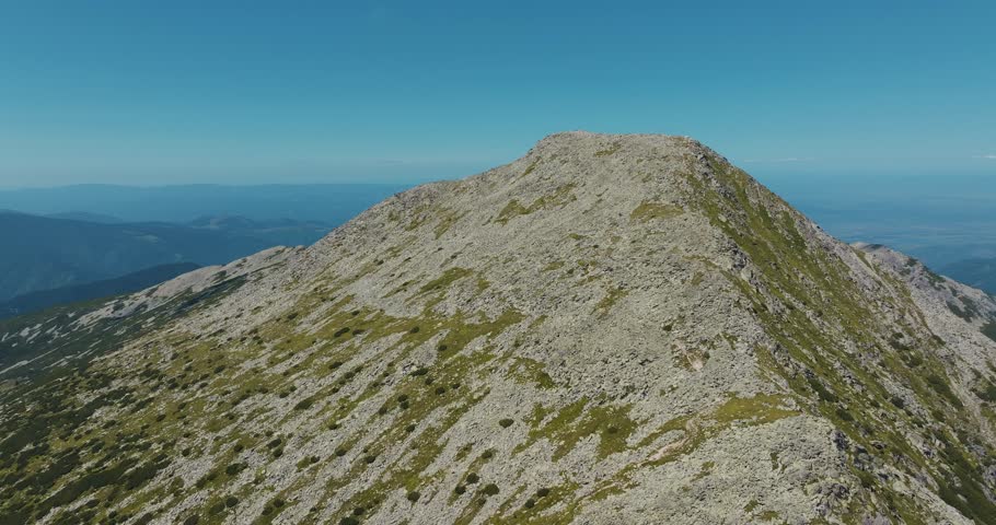 Rocky mountain tall peak on a clear sunny day with blue skies and vibrant green grass, rising drone shot