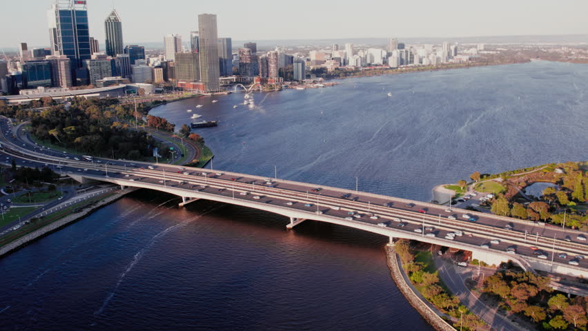 Aerial view of a vibrant city skyline of Perth, Western Australia overlooks a busy bridge traversing a river during sunset, showcasing urban life and traffic.