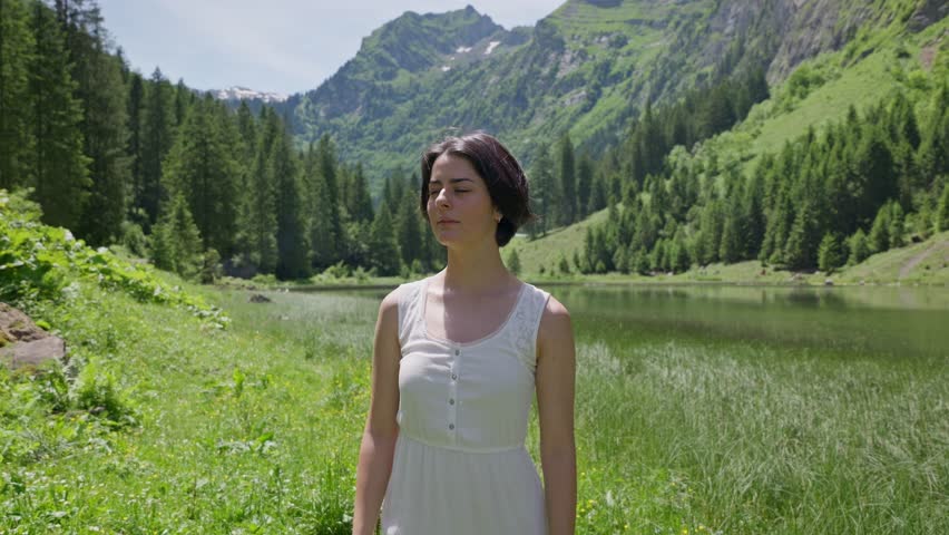 Close up shot of pretty woman with brunette short hairs looking into camera. Beautiful idyllic landscape in swiss mountains. Sunny summer day with grass, trees and lake. Dolly shot.