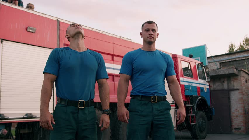 Two muscular rescuers at fire station take off their t-shirts wet from rain. Slow motion.