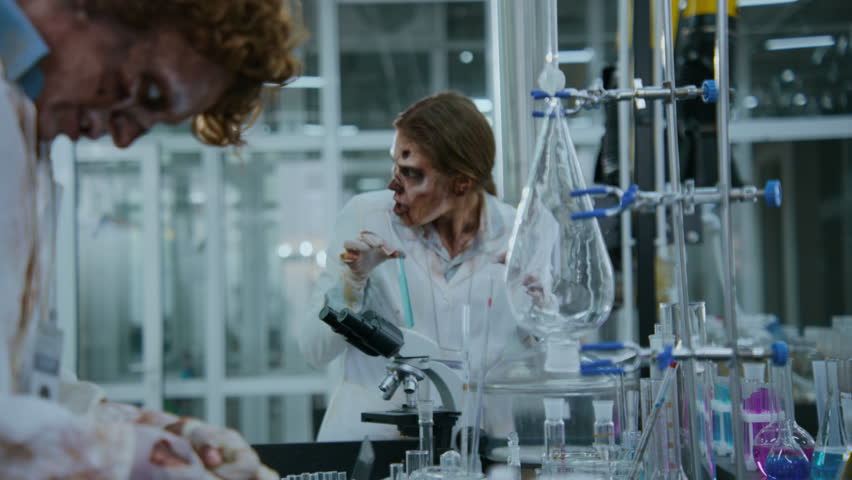 Medium shot of Caucasian female undead laboratory assistant with rotting flesh is studying blue liquid in test tube, watching male corpse colleague, who is mixing cocktail of toxic chemicals in jar