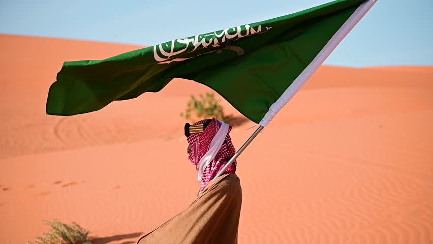 Amidst the golden desert, a Saudi man in traditional attire waves the national Saudi flag celebrating National and Founding Day with pride a stunning symbol of heritage unity, and the Kingdom's legacy