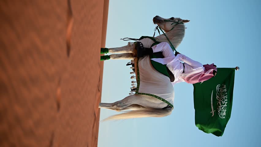 Saudi heritage in motion, a rider in traditional attire proudly waves the Saudi flag while on horseback, celebrating National and Founding Day under a vibrant sky. Arabic horse. Vertical video