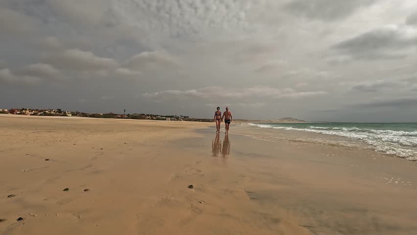 Couple in swimwear walks hand in hand along peaceful, sandy beach of Boa Vista island with cloudy sky overhead, creating serene and intimate moment by the ocean, Cape Verde