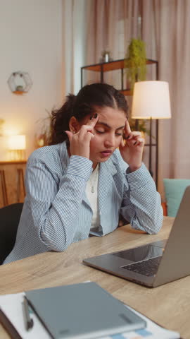 Tired ill Indian woman suffering from headache problem tension and migraine stress at home office desk workplace. Pretty freelancer broker worker girl works on laptop netbook. Overworking. Vertical.