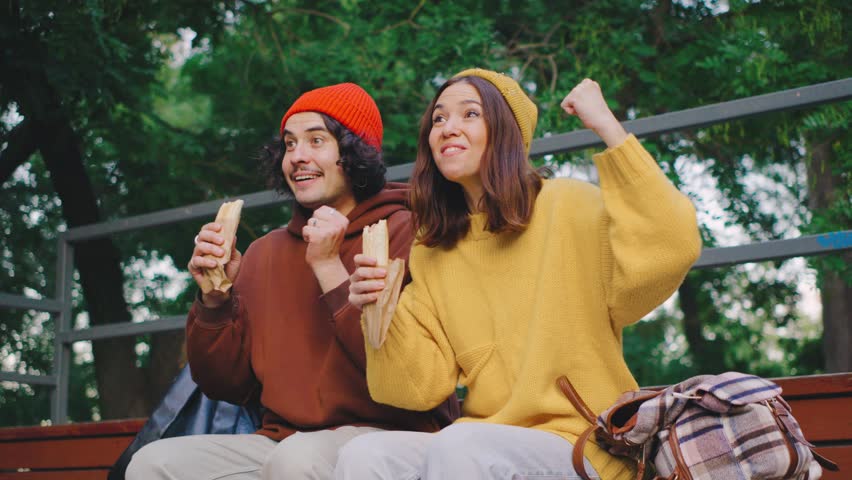 A couple sits on a park bleacher, eating hot dogs and enthusiastically cheering for their team. Their bright outfits match their joyful energy and excitement