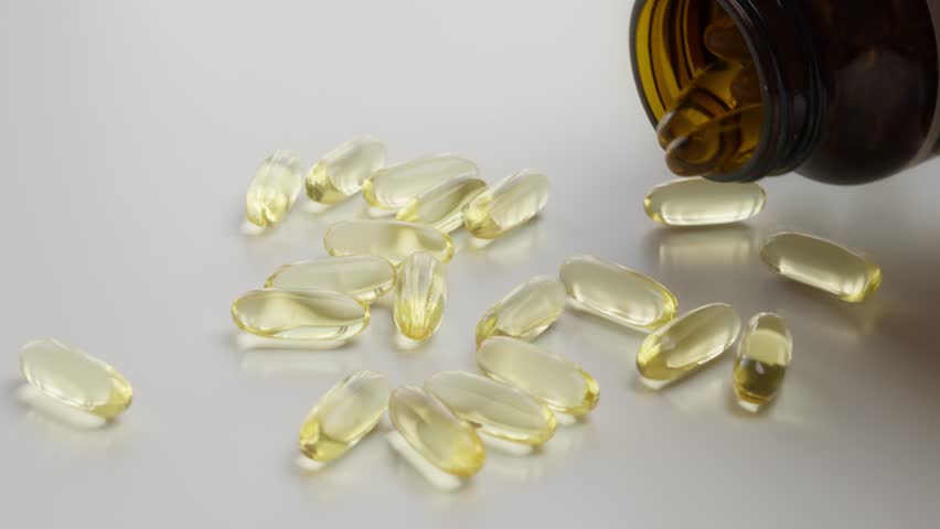 Close-up of yellow capsules spilling from a dark bottle onto a white surface. Ideal for concepts related to health, vitamins, supplements, medicine, and nutrition.