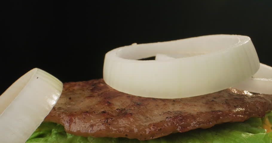 Delicious big hamburger preparation. Onion rings fall on bun with grilled BBQ patty mustard and lettuce. Party outdoors, picnic, fast food. Close-up slow motion