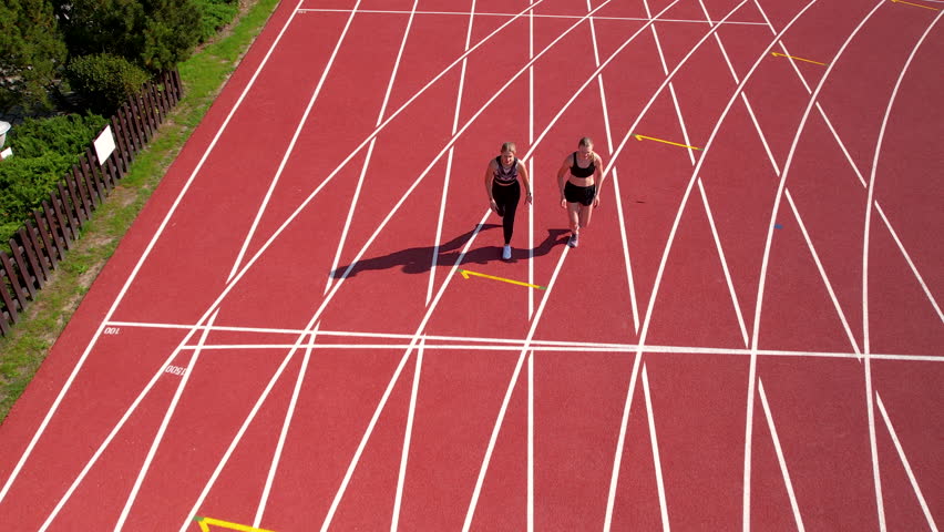 Two female athletes jogging side by side on a red running track, shadows casting towards lanes, surrounded by greenery