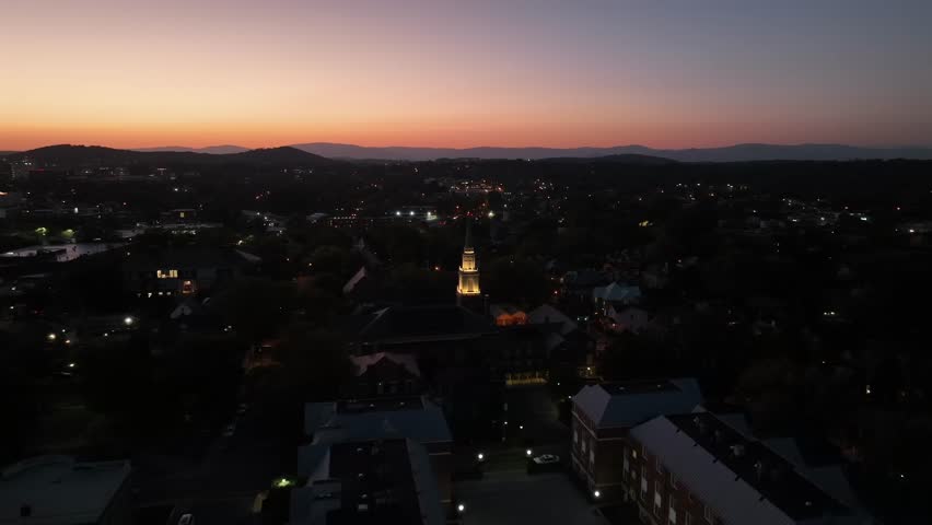 Aerial orbit shot of lighting church in dark american city after golden sunset. Dusk scene in small town with clear sky and mountain range silhouette. Wide shot.