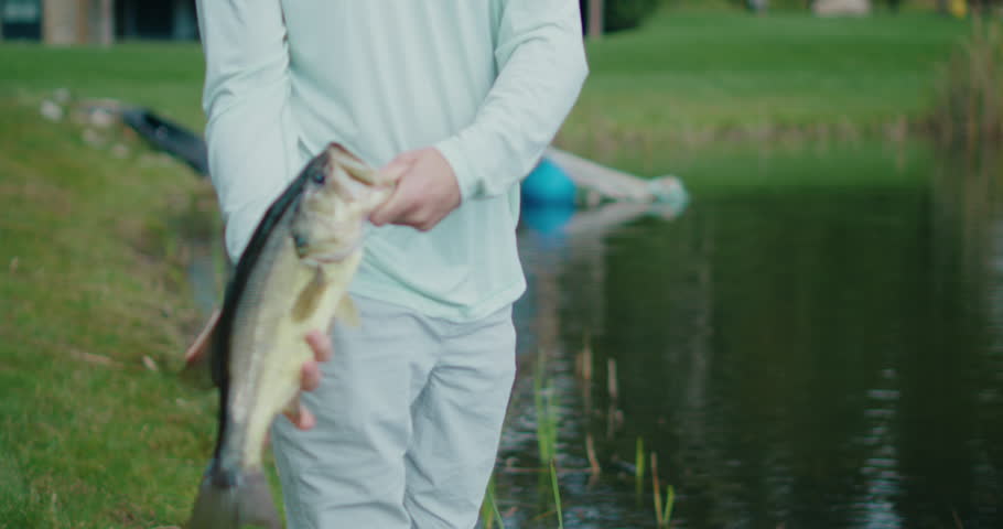 A man proudly holding a big largemouth bass he just caught while standing next to a tranquil pond. The camera moves slowly,