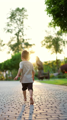 Little barefoot baby boy in t-shirt and shorts runs by the park. Kid running happily by the paved alley at sunset. Vertical video.