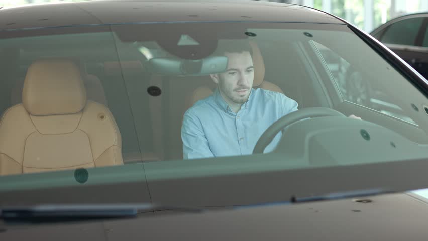 Camera view through car windshield of Caucasian man. Young male in shirt comfortably sitting in transport. Spinning steering wheel with hands while imagining driving new car. Customer of auto salon.