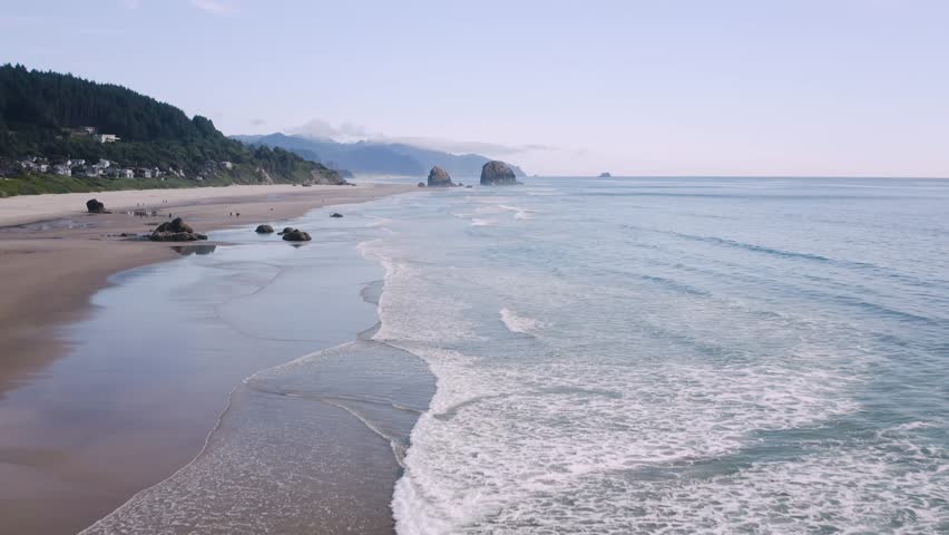 Low tide waves hit the gold sand Cannon Beach on the Oregon Coast. Seastack rock formation on the horizon. Slightly above dolly in drone flight