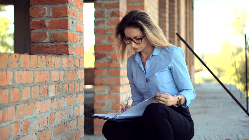 Woman Reviewing Documents at Construction Site. woman in a blue shirt sits on a ledge, studying documents at an unfinished building site. She appears focused and diligent, surrounded by brick walls. 