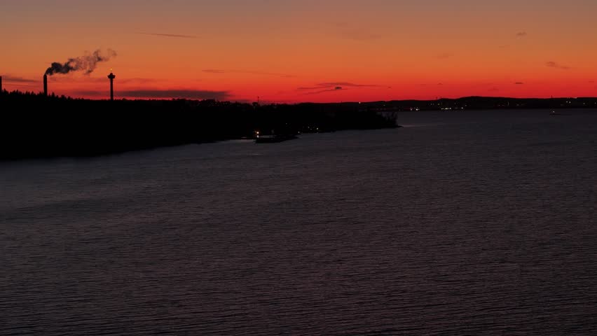 Rotating flight over a lake with dark forested shore and the sunset colours in the horizon behind silhoutte of buildings of Tampere, Finland