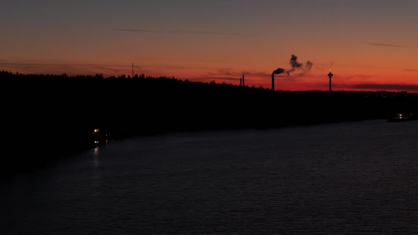 Sideways flight to the left over a lake with dark forested shore and the sunset colours in the horizon behind silhoutte of buildings of Tampere, Finland