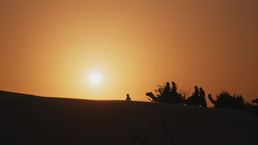 Silhouette of tourists riding camels on top of a dune in a safari desert tour during sunset